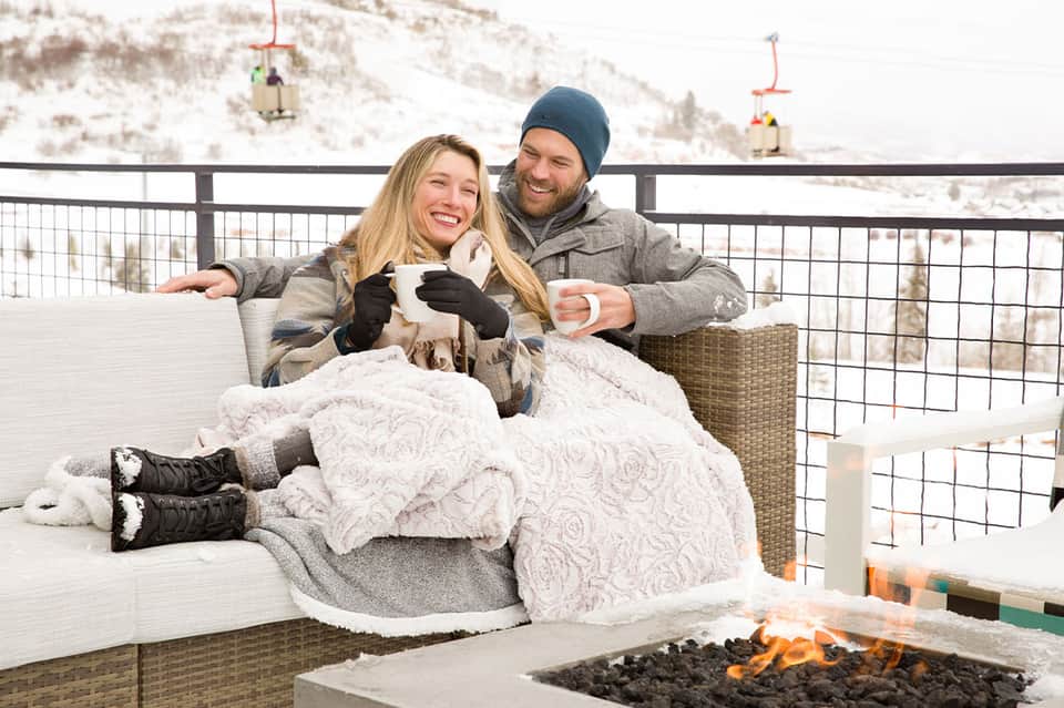 Couple wrapped in blankets enjoying hot drinks on a snowy mountain overlook with ski lifts visible