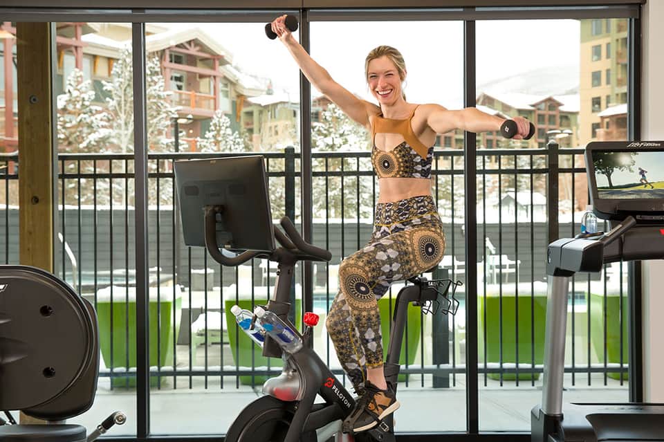 Woman exercising on stationary bike in modern gym with panoramic windows overlooking snowy mountains