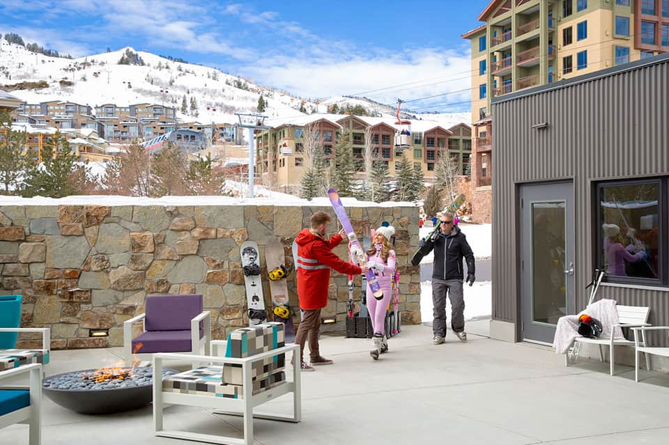 Mountain ski resort terrace with fire pit, modern furniture, snow-covered slopes, and residential buildings