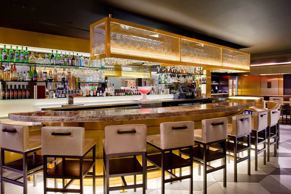 Modern bar interior with amber lighting, beige bar stools, and fully stocked shelves behind counter