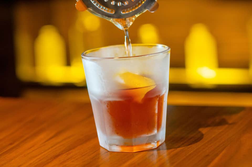 Bartender pouring whiskey into a glass with ice and an orange slice on a wooden bar counter