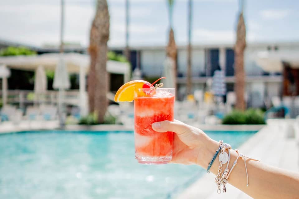 Hand holding coral cocktail with orange slice by poolside lounge area