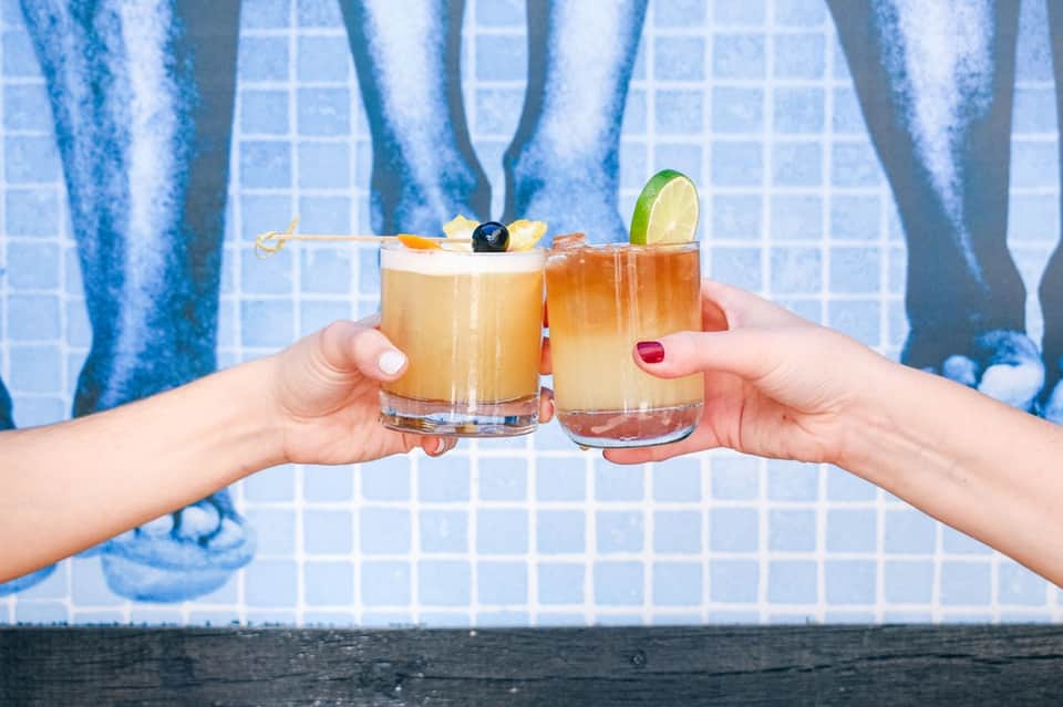 Two hands holding tropical cocktails by pool with lime garnish and olives