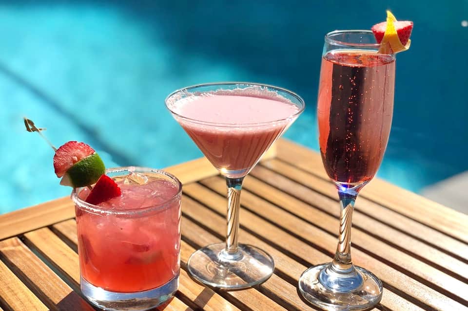 Three colorful cocktails with garnishes on wooden deck by bright blue pool