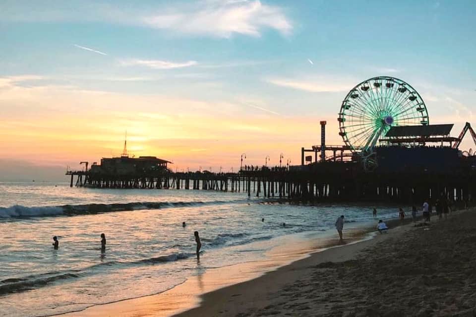 Beach scene at sunset with pier, Ferris wheel, and people swimming in the ocean