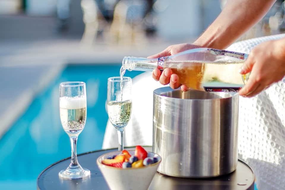 Hands pouring champagne by a poolside with glasses and fruit bowl