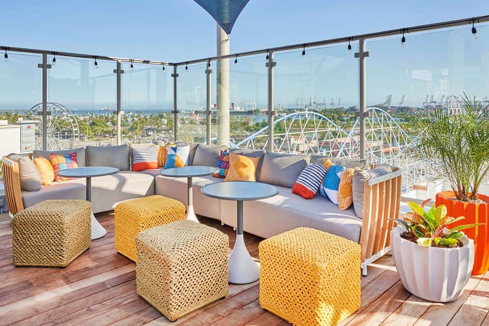 Rooftop terrace with gray seating, woven yellow poufs, colorful pillows, and amusement park views