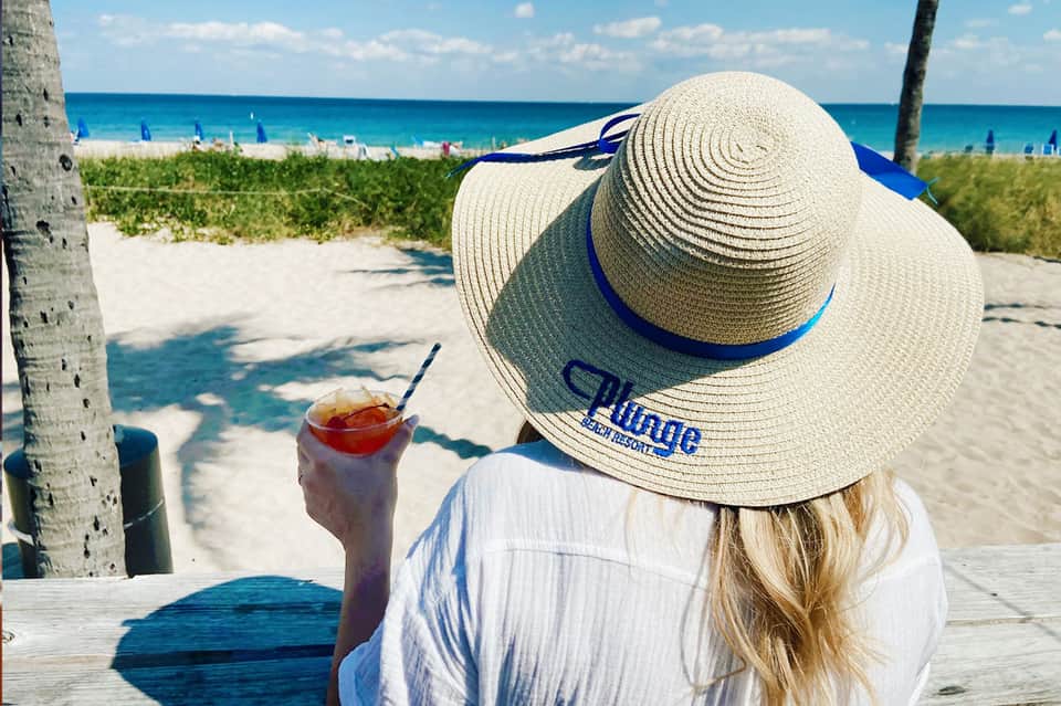 Woman in wide-brimmed hat holding tropical drink overlooking beach