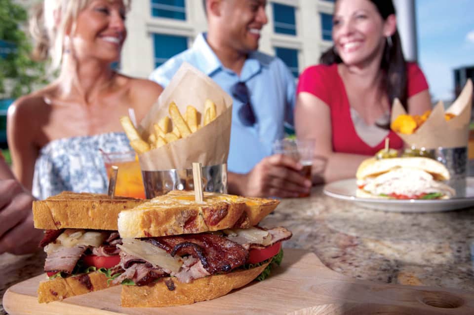 Group dining outdoors with gourmet sandwich and french fries on table