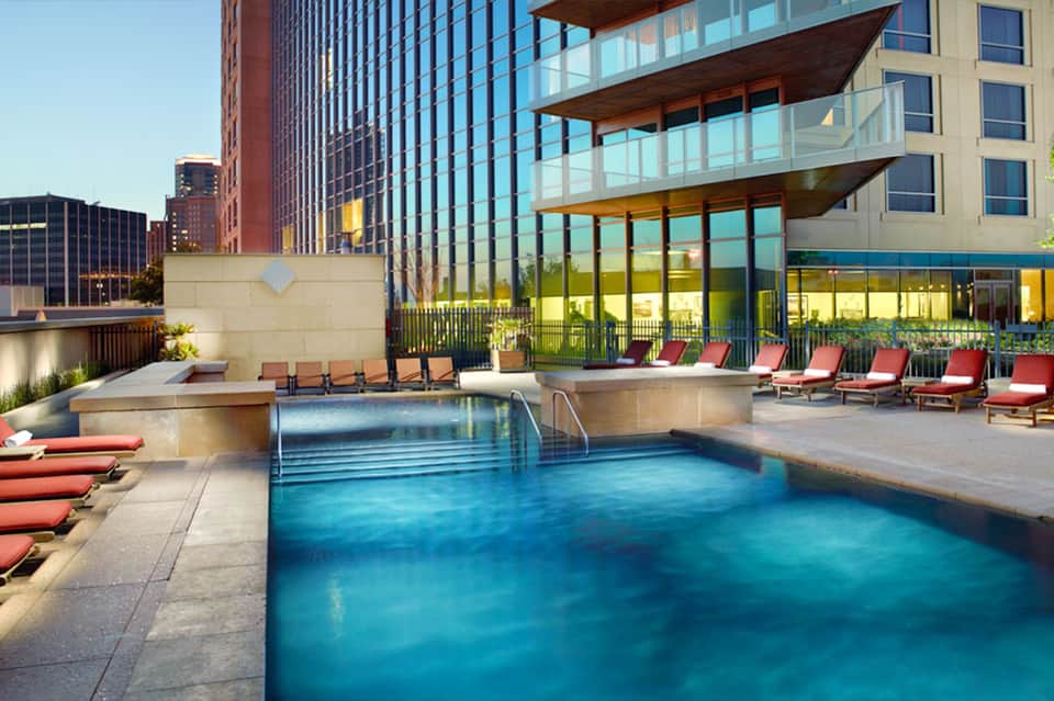 Modern rooftop pool with turquoise water, red lounge chairs, and city skyline with tall buildings