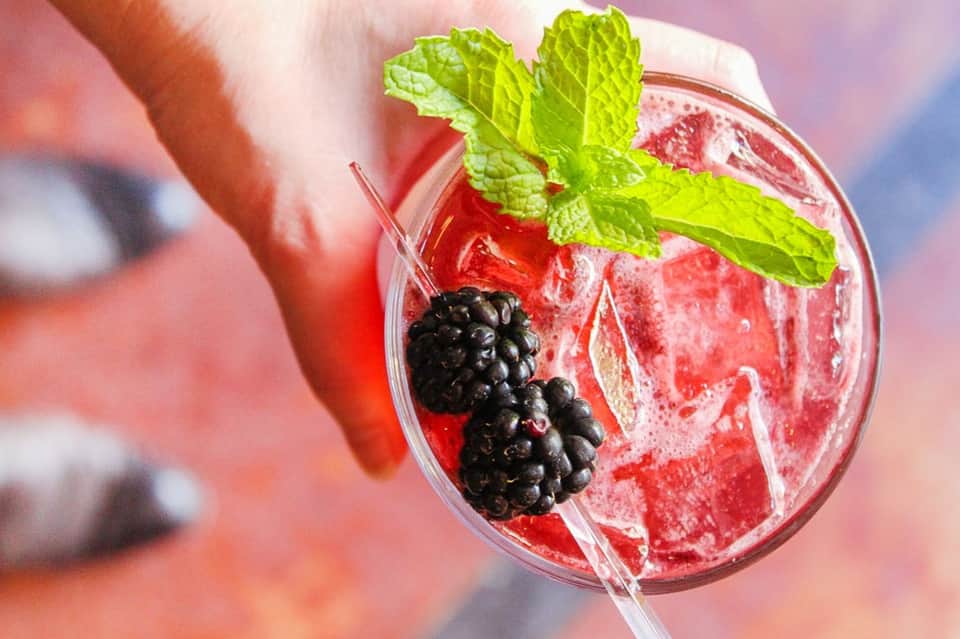 Red cocktail with ice, fresh blackberries, mint leaves, and straw in clear glass