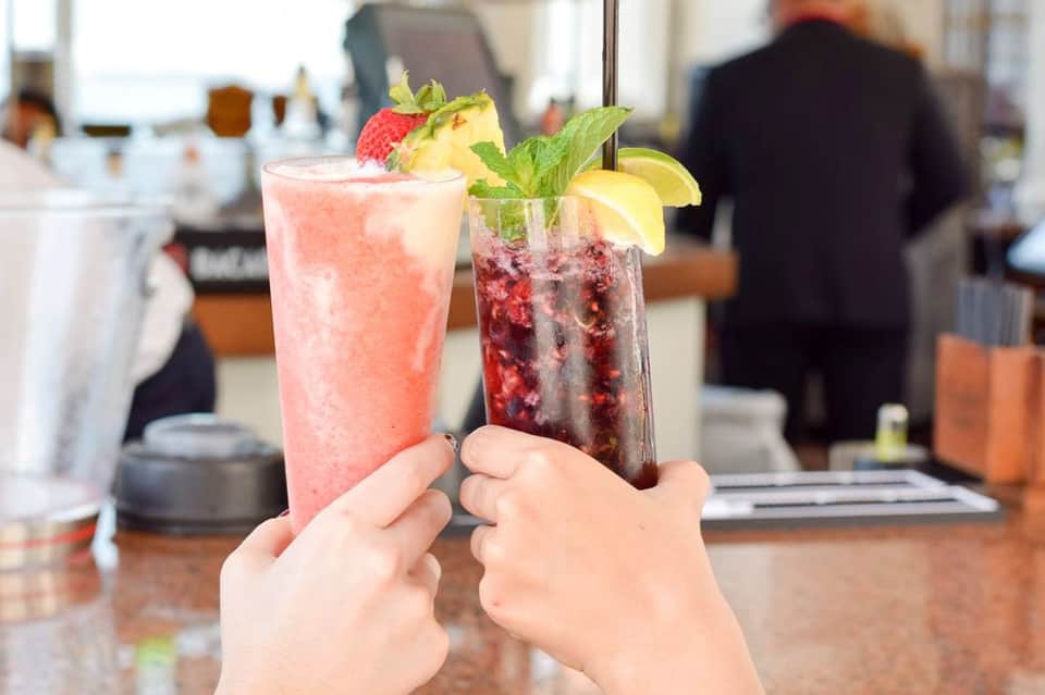 Two colorful cocktails held in hands at bar counter - pink drink and dark berry beverage with fruit garnishes