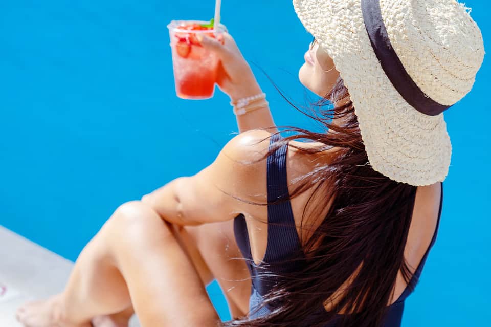 Woman in straw hat holding tropical cocktail against clear blue sky, wearing dark swimsuit