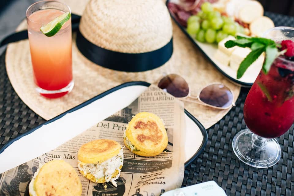 Tropical poolside dining setup with cocktails, pastries, straw hat, sunglasses, and fresh fruit