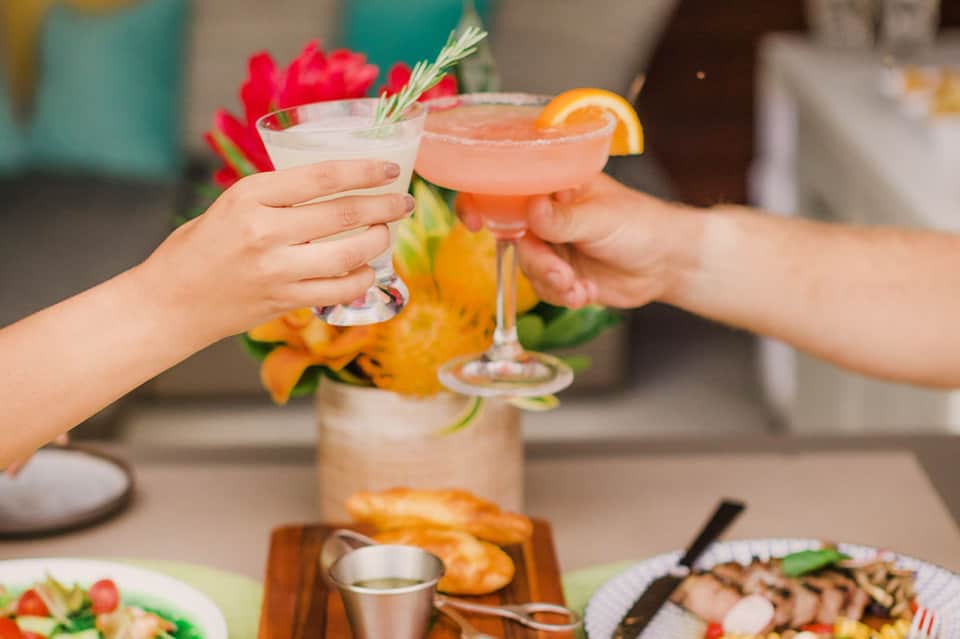 Two cocktails clinking together poolside with fresh flowers and appetizers nearby