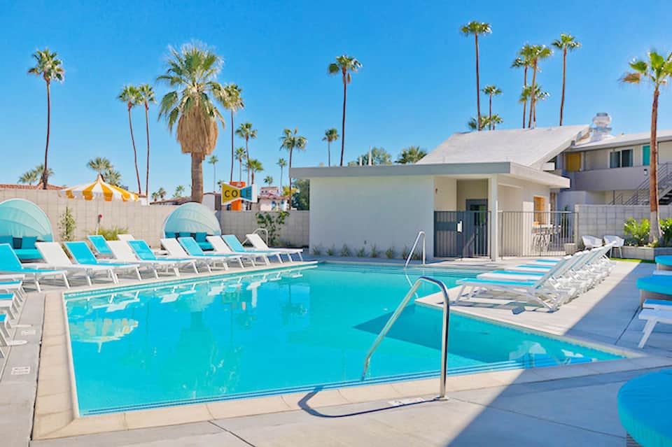 Bright turquoise swimming pool with lounge chairs, umbrellas, and palm trees under blue sky