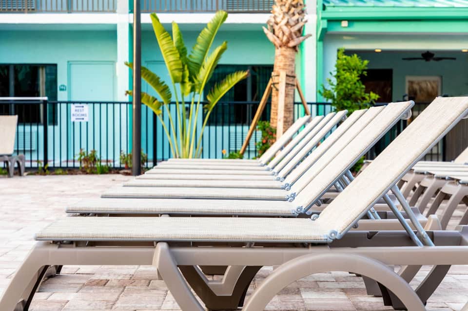 Row of white lounge chairs on a sunny patio with turquoise building and palm trees