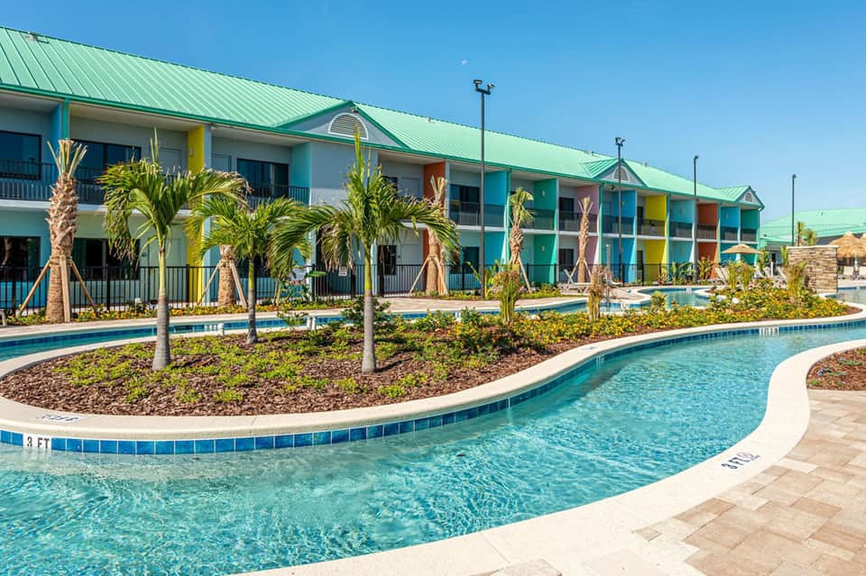Colorful resort buildings with green roof reflected in curved lazy river pool surrounded by palm trees