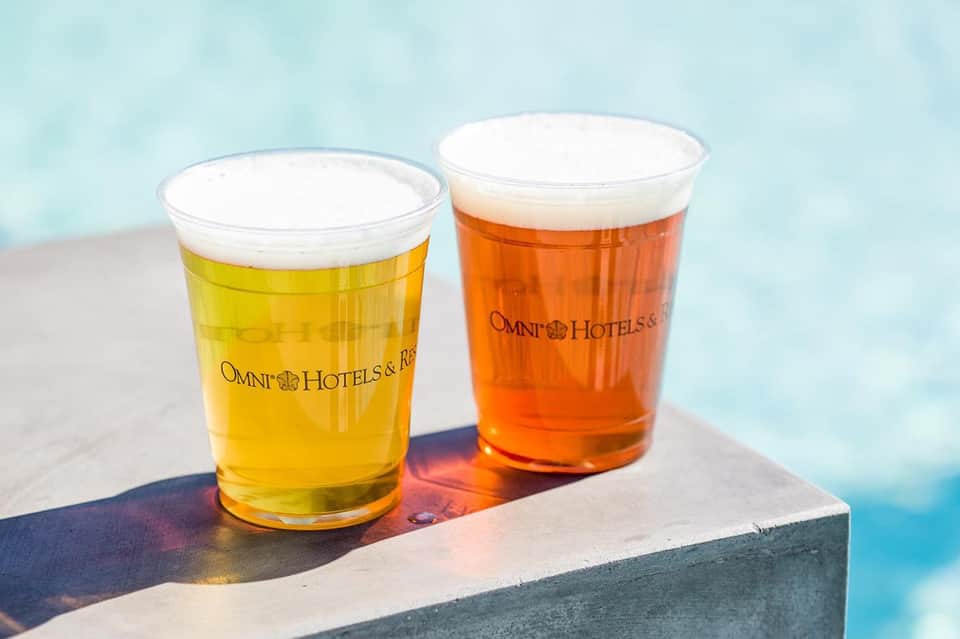 Two Omni Hotels branded beers with white foam heads on a poolside ledge against blue water