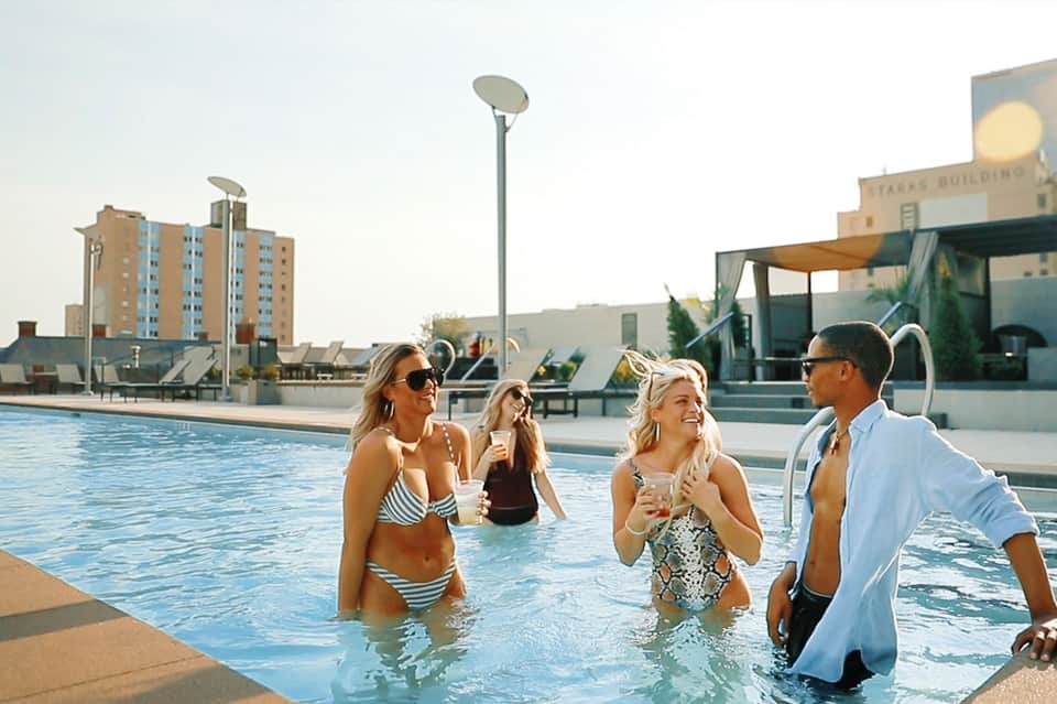 Group of four people enjoying drinks in an outdoor rooftop swimming pool with city buildings in the background