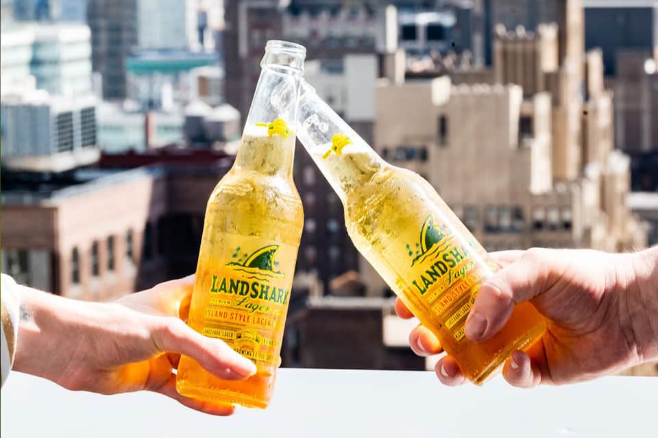 Two hands toasting with Landshark beer bottles against city skyline backdrop