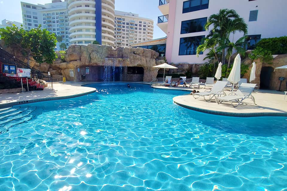 Resort pool with rock waterfall feature, lounge chairs, umbrellas, and high-rise hotel buildings in background