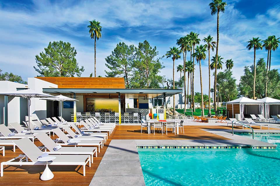 Modern resort pool area with turquoise water, white lounge chairs, wooden deck, and palm trees under blue sky