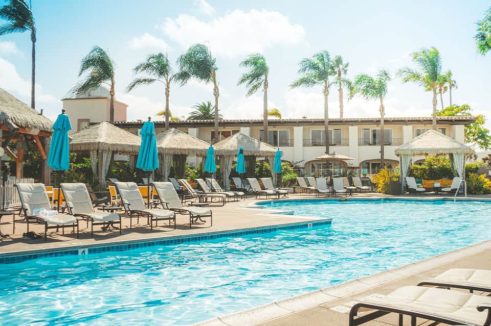 Resort pool with turquoise umbrellas, lounge chairs, palm trees, and Spanish colonial buildings