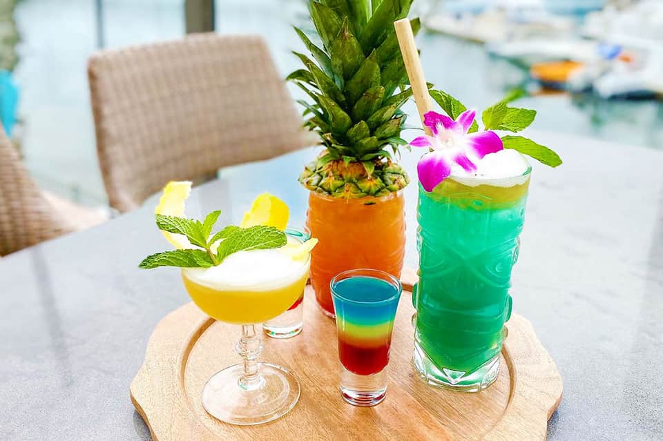 Three tropical cocktails on wooden board featuring pineapple, colorful drinks, and floral garnishes