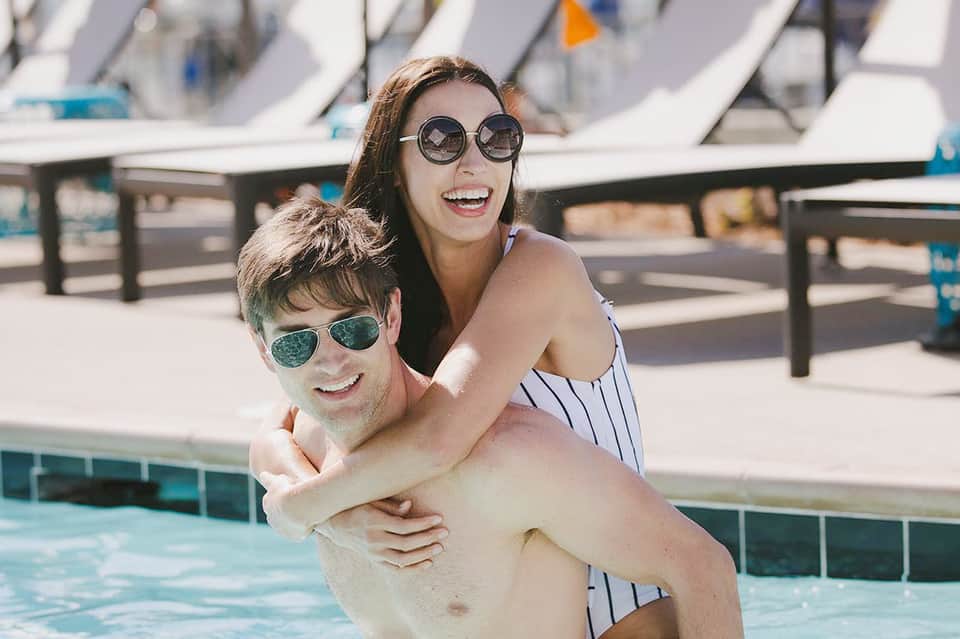 Man and woman smiling by pool, woman wearing round sunglasses and striped swimsuit