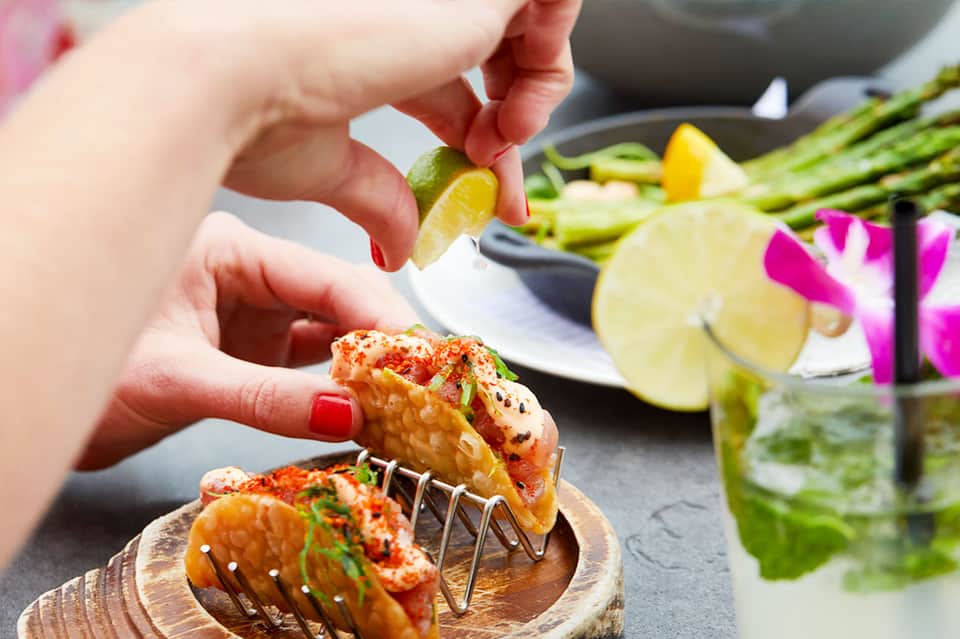 Hands holding grilled shrimp with lime, garnished with spices near a cocktail with flowers