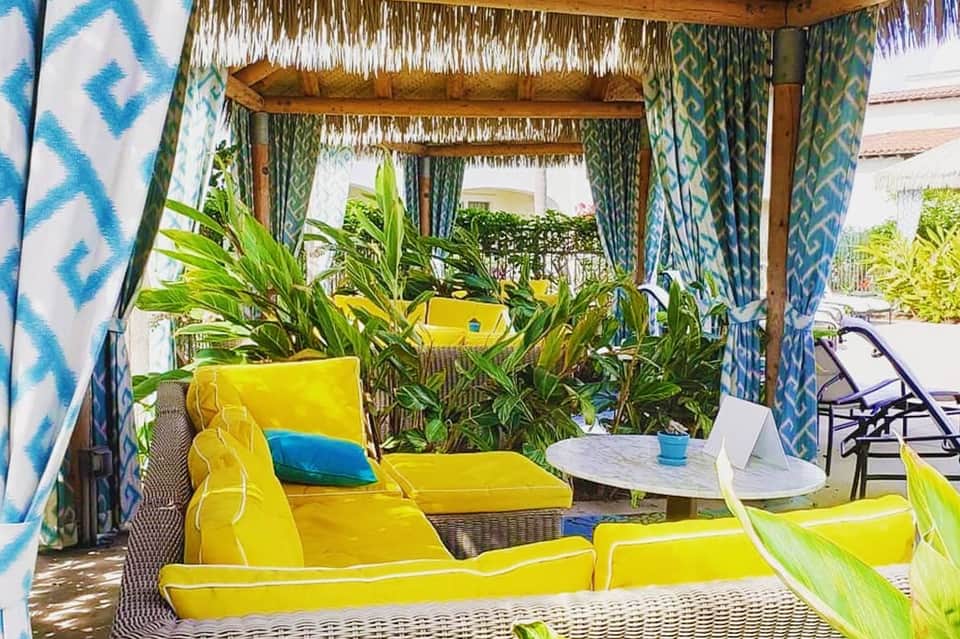 Tropical cabana with yellow cushioned seating, patterned blue curtains, plants, and white shade structure
