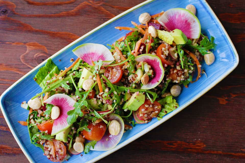 Blue rectangular plate with quinoa salad, radish slices, leafy greens, nuts, and colorful vegetables on wooden surface