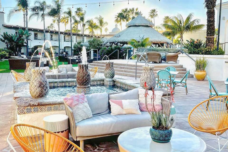 Luxury resort courtyard with lounge seating, decorative pineapple fountains, and thatched-roof structures surrounded by palm trees