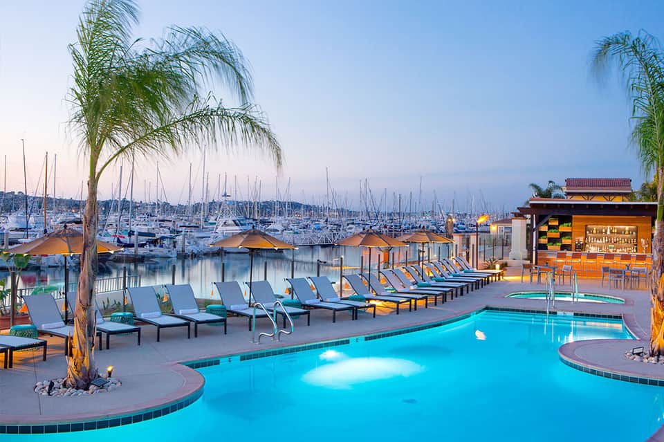Illuminated poolside lounge with blue water, palm trees, and sailboats docked in the marina at dusk
