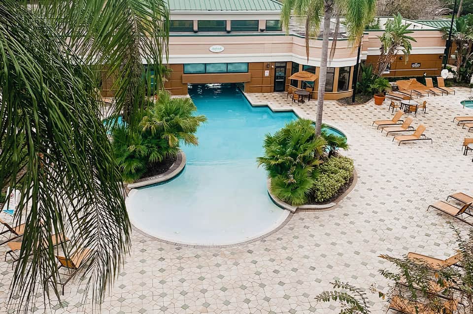 Tropical resort pool with curved edges, palm landscaping, lounge chairs, and modern building with glass windows