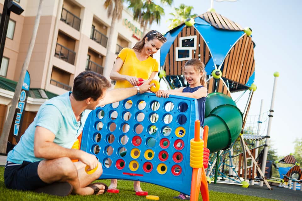 Family playing Connect Four game on lawn with playground equipment and resort buildings