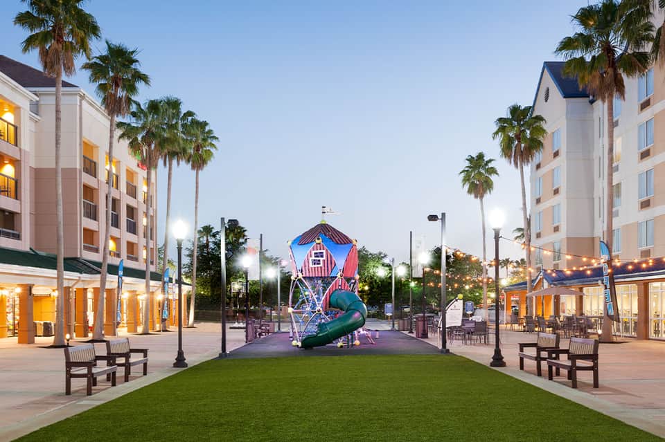 Hotel courtyard at dusk with illuminated buildings, palm trees, and colorful playground sculpture