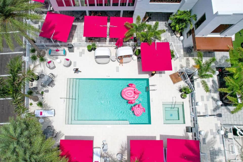 Aerial view of resort pool deck with turquoise water, pink umbrellas, lounge chairs, and tropical landscaping