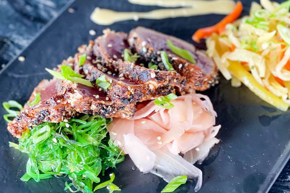 Seared tuna slices with sesame seeds, ginger, and green onions on dark plate with noodles