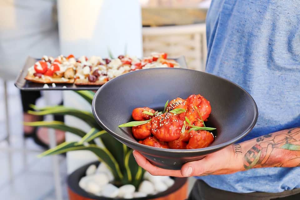 Hand holding bowl of glazed strawberries with appetizers blurred in background