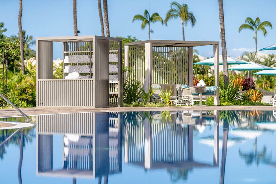 Modern poolside cabana with vertical slat design reflected in calm water, surrounded by palm trees