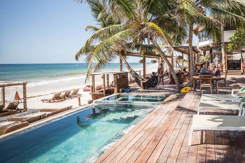 Beachfront beach club with wooden deck, infinity pool, palm trees, and ocean view with loungers