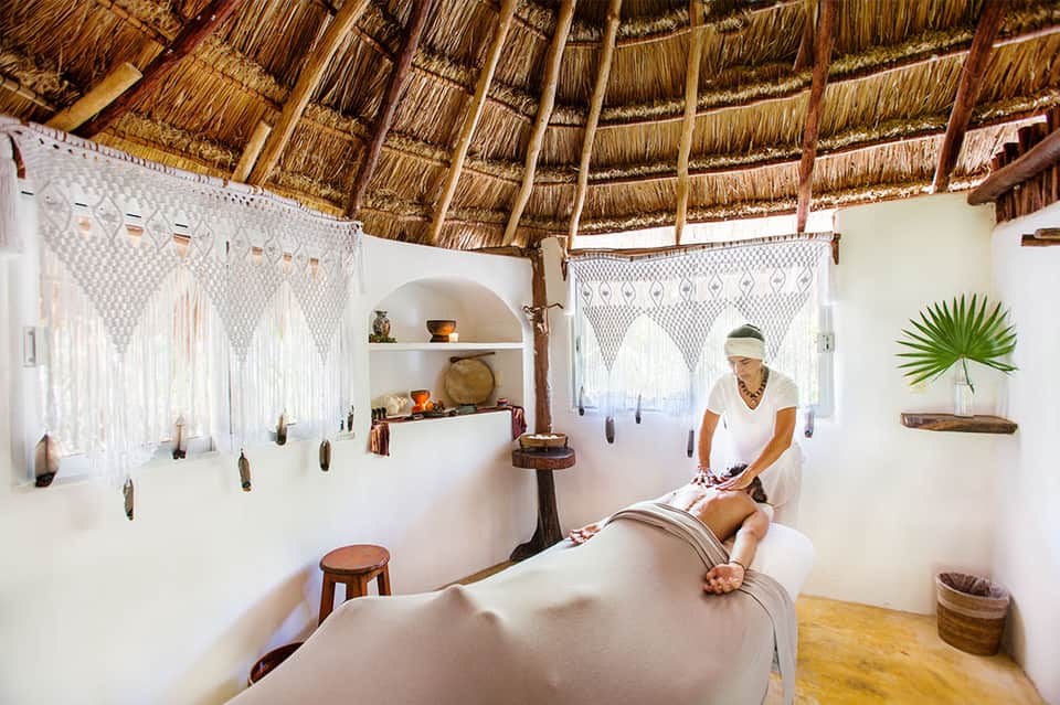 Spa treatment room with thatched roof, white walls, and therapist performing massage on client