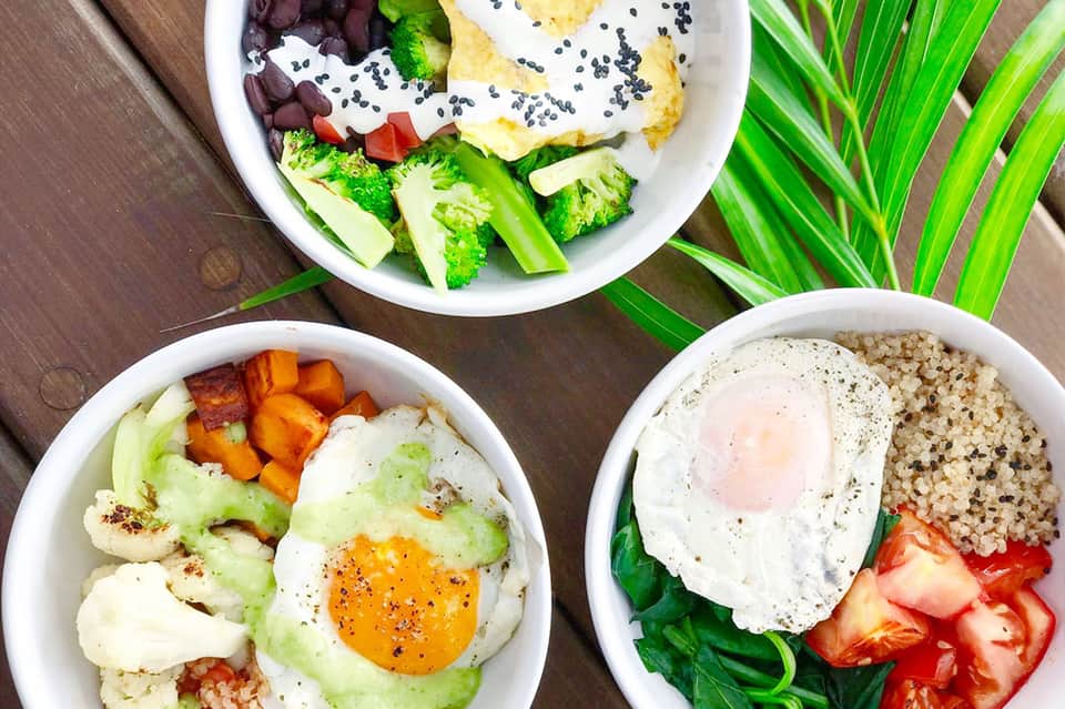 Three white bowls of healthy meal prep with eggs, vegetables, grains, and fresh greens on wooden surface