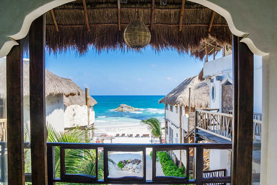 Beachfront bungalow view framed by thatched roof, overlooking turquoise ocean and white sand beach