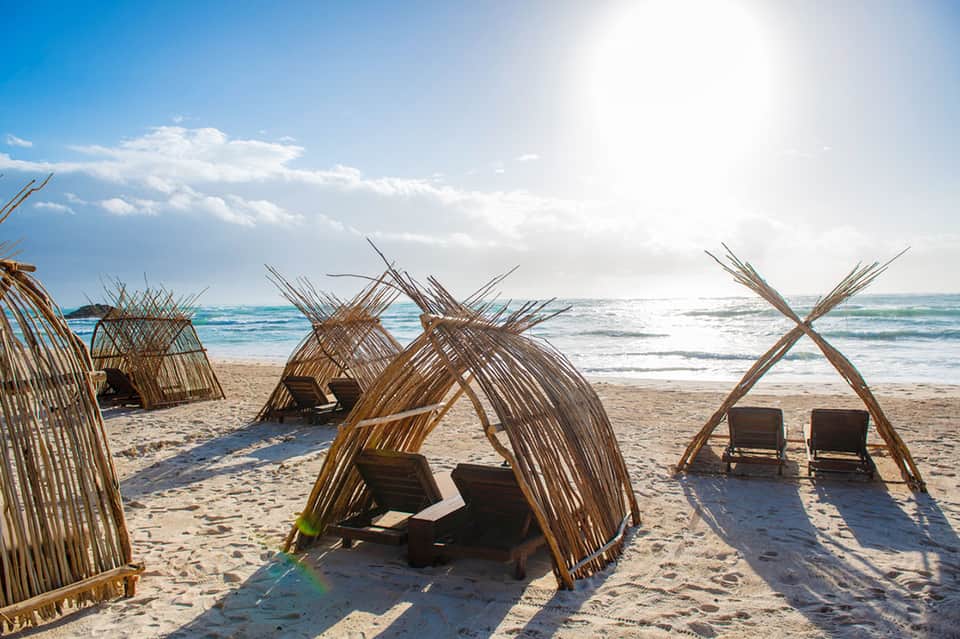 Beach scene with woven wooden tent structures and lounge chairs on sand facing the ocean