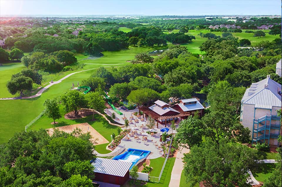 Aerial view of sprawling resort with pool, recreational facilities, and manicured green grounds surrounded by trees