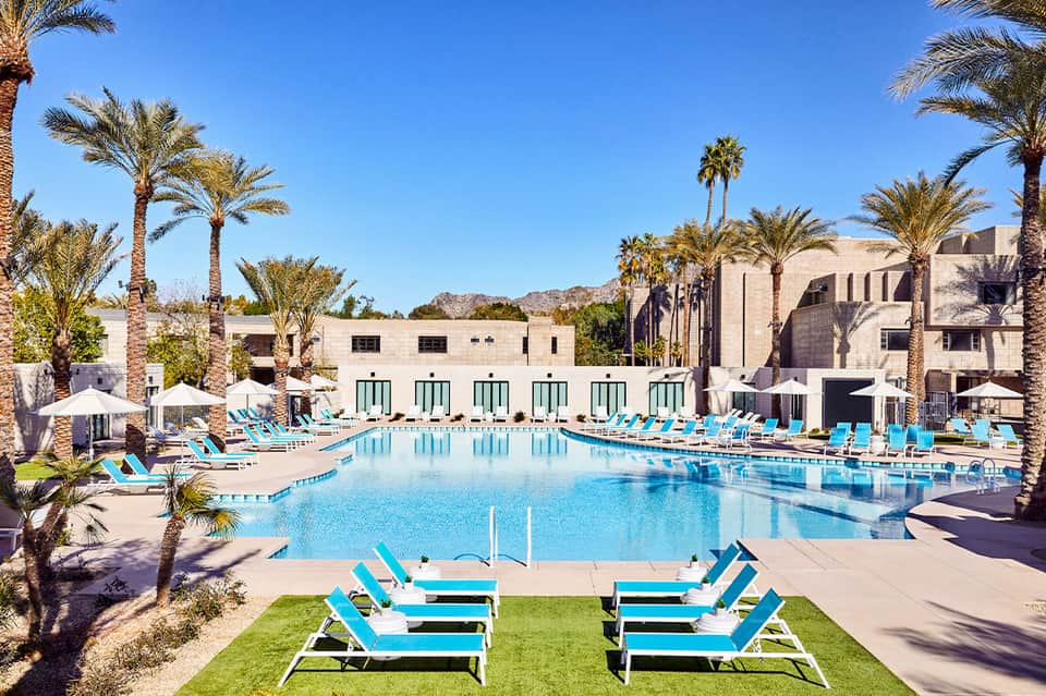 Large resort pool with turquoise loungers, palm trees, and modern white buildings under clear blue sky