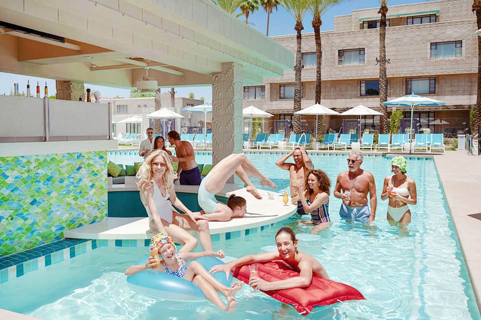 Outdoor pool with people enjoying water activities, float toys, covered cabana area, and modern hotel architecture